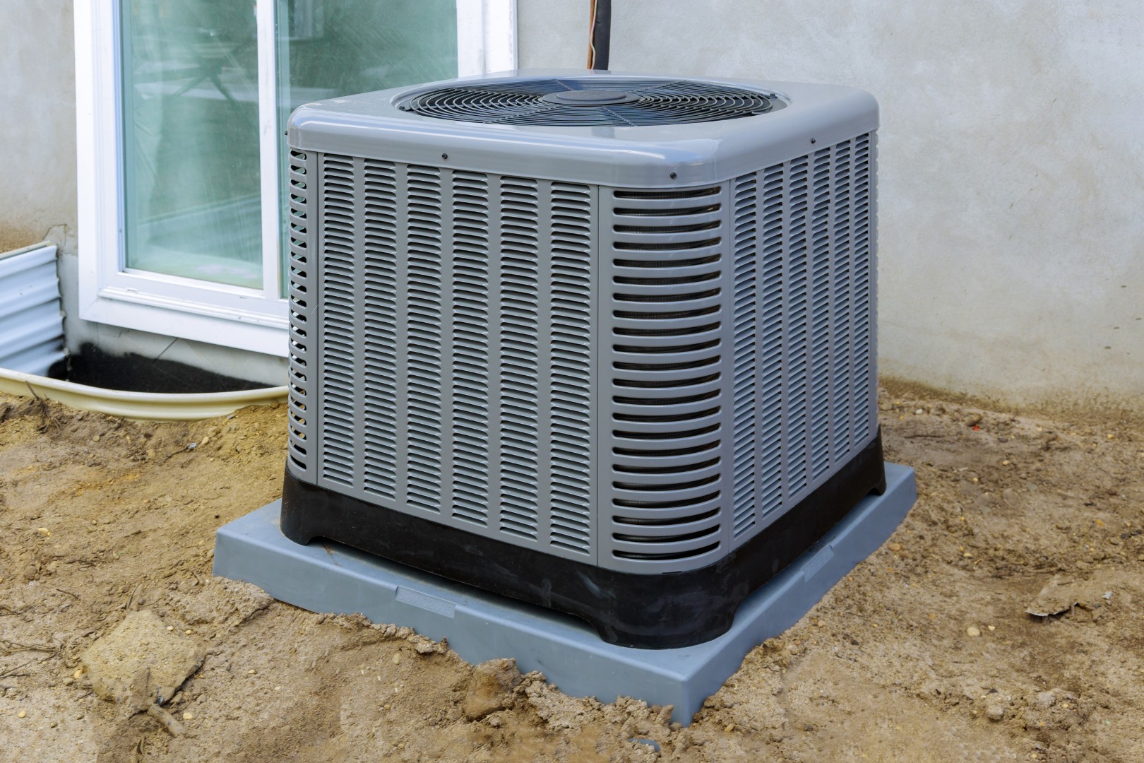 A central air conditioning unit sits on a concrete pad outside a building near a window, surrounded by bare soil—ready for inspection or ac repair if needed.