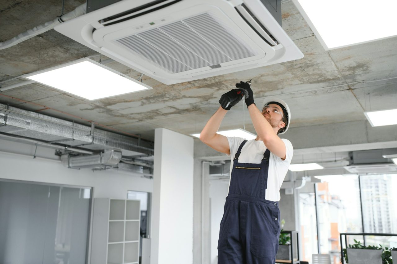 A worker in overalls and a helmet uses a tool to perform ac repair on a ceiling-mounted air conditioning unit in a modern office.